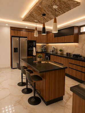 Una cocina moderna con una isla central, taburetes de bar, armarios de madera y electrodomésticos de acero inoxidable, además de iluminación colgante.