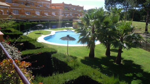 Una piscina rodeada de césped cuidado, palmeras y un edificio de varios pisos con balcones.
