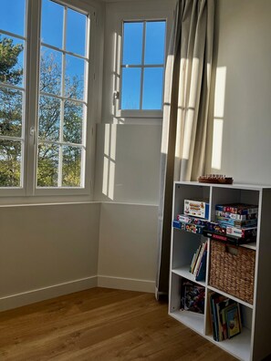 Ein Eckzimmer mit Holzboden, einem weißen Bücherregal mit Gesellschaftsspielen und einem fenster mit Vorhängen.