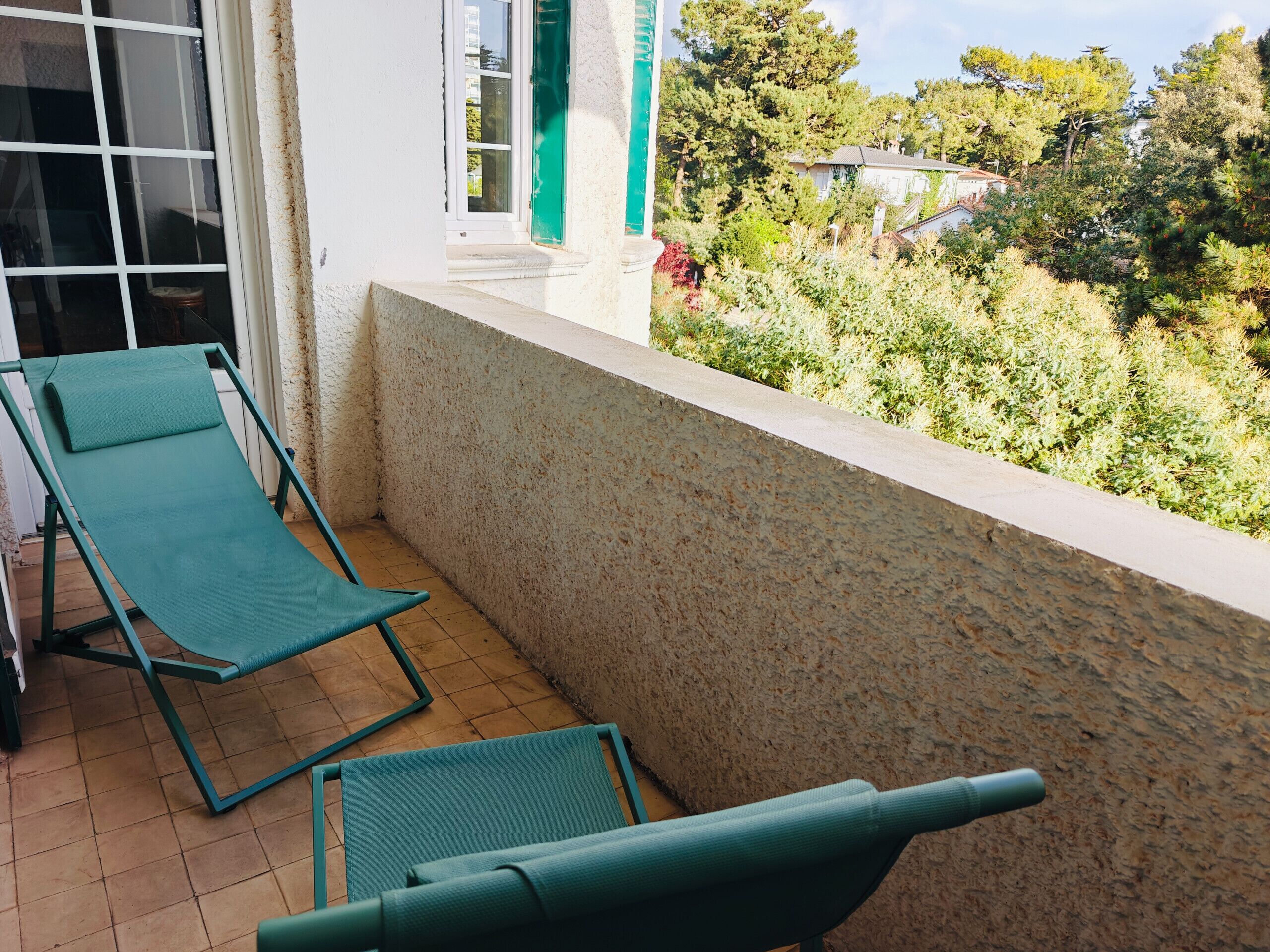 Un balcon avec deux fauteuils de jardin verts, un mur blanc et une vue sur un jardin et des maisons.