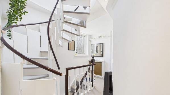 Eine Wendeltreppe mit weißen Geländern und einer dunklen Holzhandläufe, die zu einem Zwischenpodest mit Kamin und gerahmten Kunstwerken an den Wänden führt.