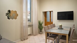 A modern living room with a dining area, a large window with curtains, a flat-screen TV mounted on the wall, and a potted plant. - Toledo Rooftop | Panoramic Terrace | Grandi Residenze Napoli (Napoli)