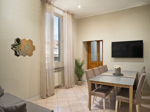 Ein modernes Wohnzimmer mit Essbereich, einem großen Fenster mit Vorhängen, einem flachbildfernseher an der Wand und einer Topfpflanze.