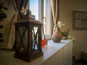 Ein Zimmer mit Fenster, einer Blumenvase, einer Kerze und einem an der Wand hängenden Kunstwerk.