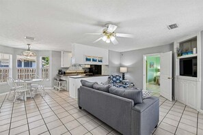 Eine moderne Küche mit weißen Schränken, ein Essbereich mit weißem Tisch und Stühlen, ein graues Sofa und ein Deckenventilator.