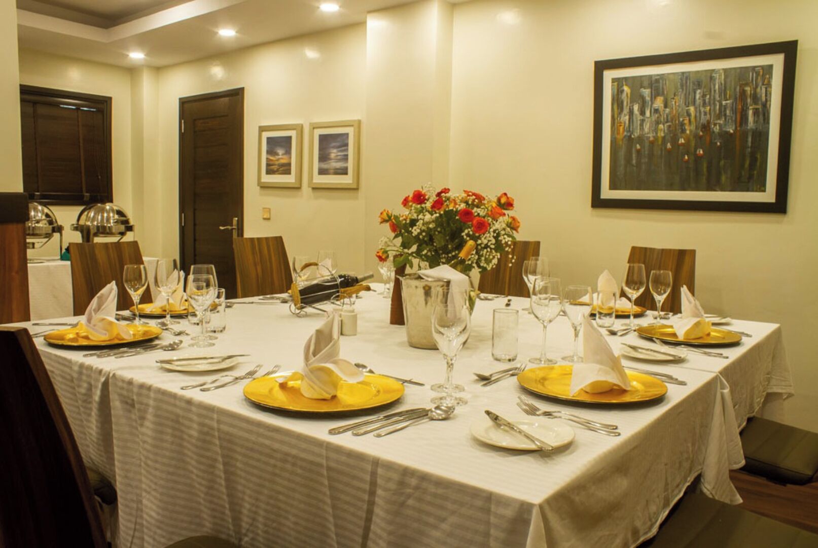 A dining room with a large round table set for a meal, framed artwork on the walls, and a window with curtains.
