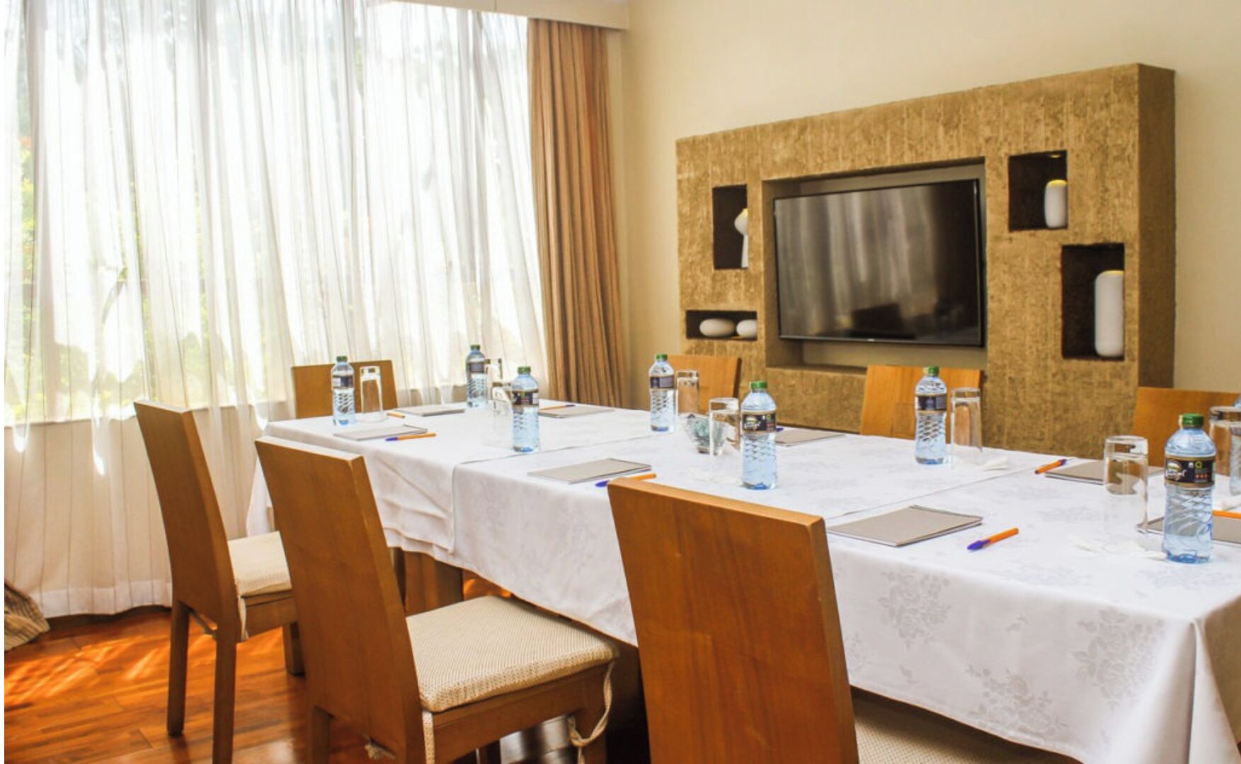 Ein Konferenzraum mit einem langen Tisch, Stühlen, Wasserflaschen, Notizblöcken, Stiften und einem an der Wand montierten Fernseher.