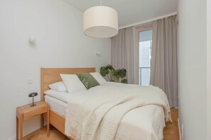 Ein Schlafzimmer mit hölzernem Kopfteil, einem Nachttisch mit Lampe, einem großen Fenster mit Vorhängen und einer Topfpflanze.