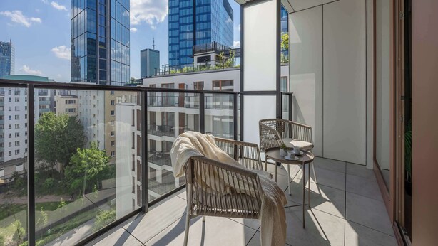 Un balcón con una silla de mimbre, una mesita y vistas de edificios altos.