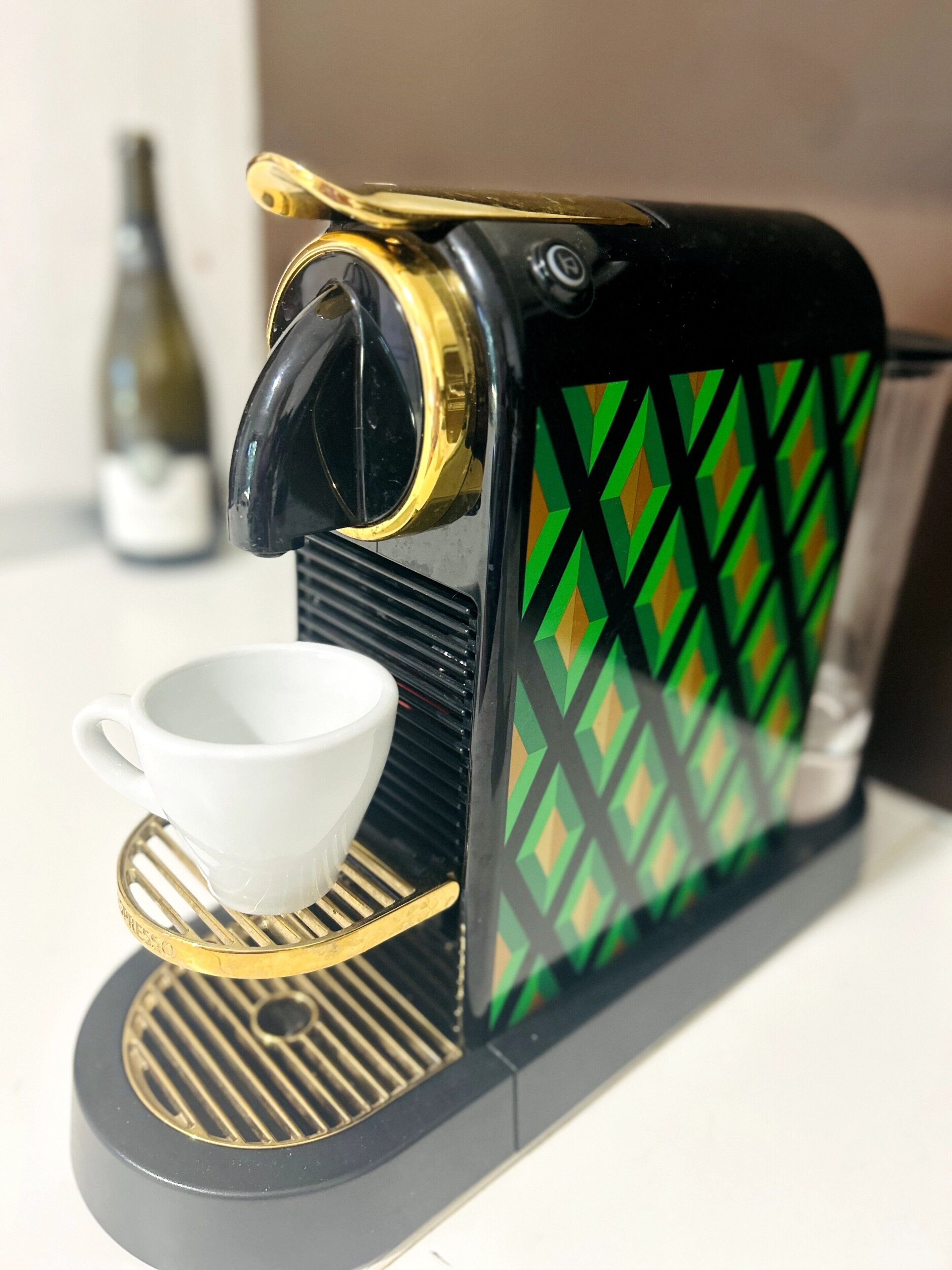 Een koffieapparaat met een kopjeshouder, een fles op de achtergrond en een decoratief patroon op het apparaat.