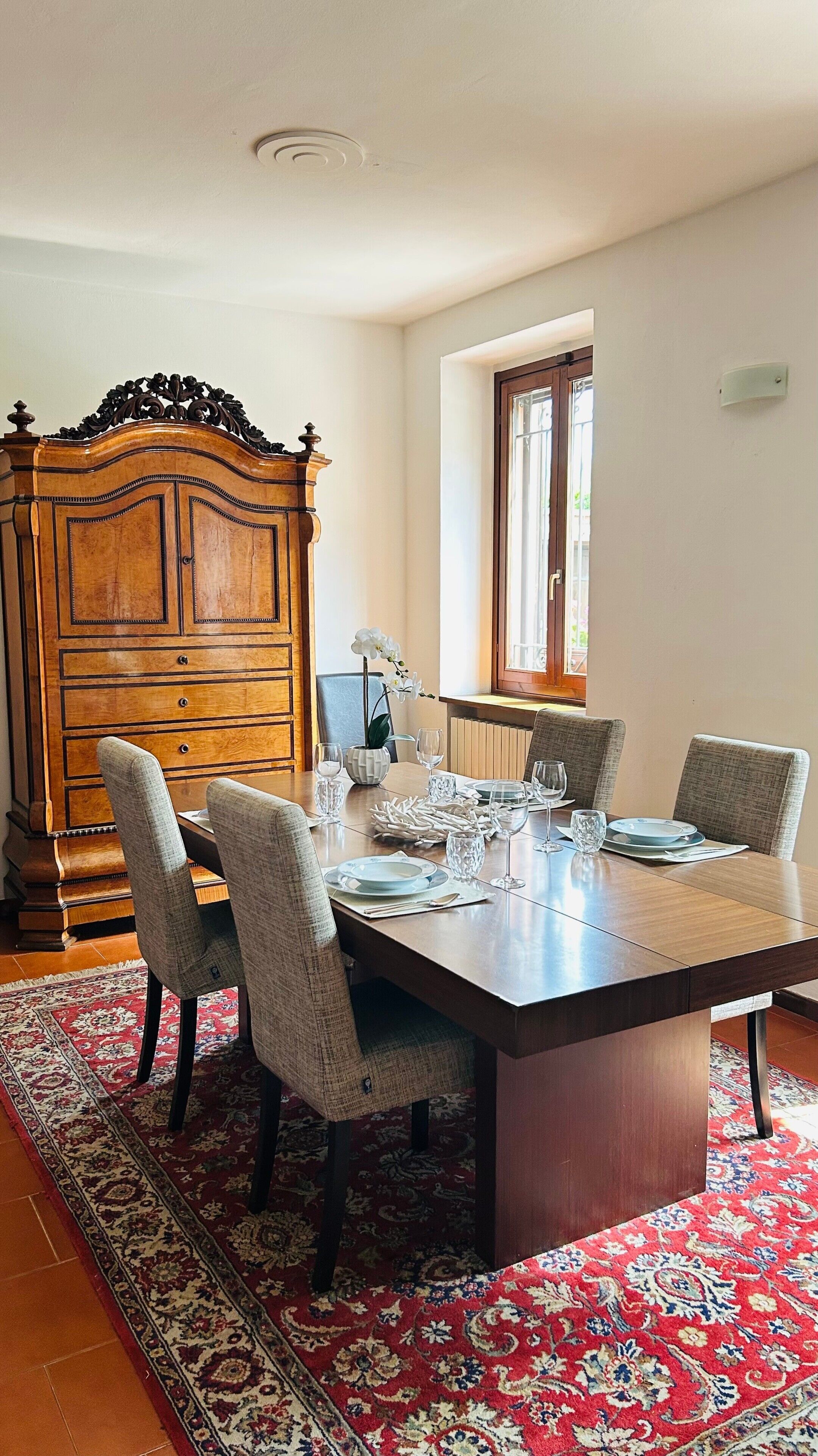 A dining room with a wooden table set for four, surrounded by chairs, a large wooden cabinet, and a patterned rug.