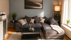 Una sala de estar moderna con un sofá gris, una mesa de centro, un cuadro enmarco con un paisaje, una lámpara de pie y una pequeña mesita con una planta.