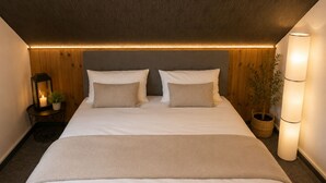 Un dormitorio con cama, dos almohadas, una mesita de noche con lámpara, una planta en maceta y una lámpara de pared.