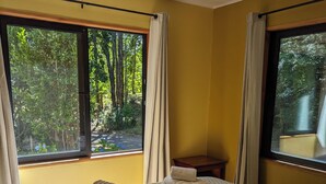 A bedroom with a bed, two windows, a nightstand, and a view of trees. - Kari Mapu Park (Pucón)