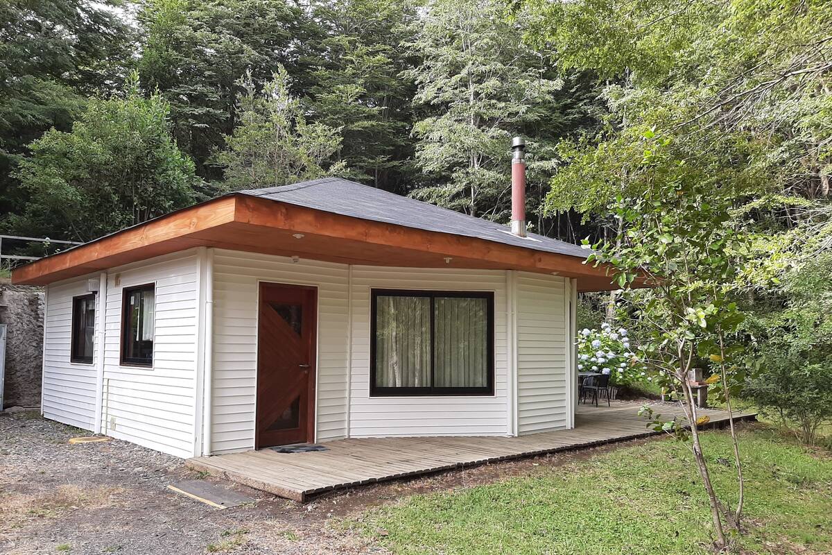 A small, single-story cabin with a wooden deck, surrounded by trees.