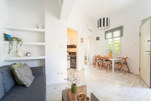 A modern living room with a sofa, a dining table with chairs, a kitchen in the background, and a door to the right. - Magic Garden House 33, near sea & center (Athens)
