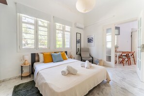 A bedroom with a bed, a nightstand, a chair, a table, and a door leading to a dining area. - Magic Garden House 33, near sea & center (Athens)