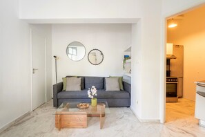 A modern living room with a grey sofa, a glass coffee table, and a kitchen visible through an open doorway. - Magic Garden House 33, near sea & center (Athens)