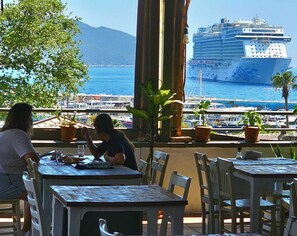 Un espace repas avec des tables et des chaises, une vue sur la mer et un navire de croisière en arrière-plan.