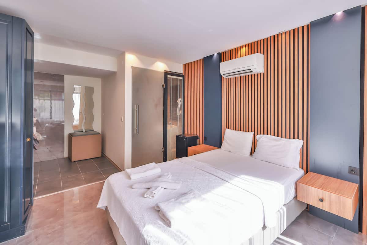 Moderni hotellihuone, jossa on suuri sänky, puupanelilla verhoiltu seinä, ilmastointilaite ja pieni yöpöytä.