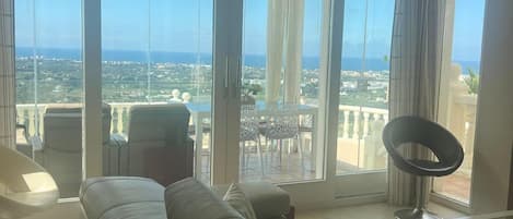 Ein großzügiges Wohnzimmer mit einer großen Couch, einem Esstisch und Blick auf Stadt und Meer durch raumhohe Fenster.