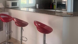 Cocina moderna con microondas, horno y encimera de granito. Hay tres taburetes rojos y un reloj en la pared.