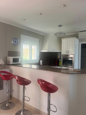 A modern kitchen with a microwave, oven, and granite countertop. There are three red bar stools and a clock on the wall. - Villa Sleeps 4 With Indoor Pool (Denia)
