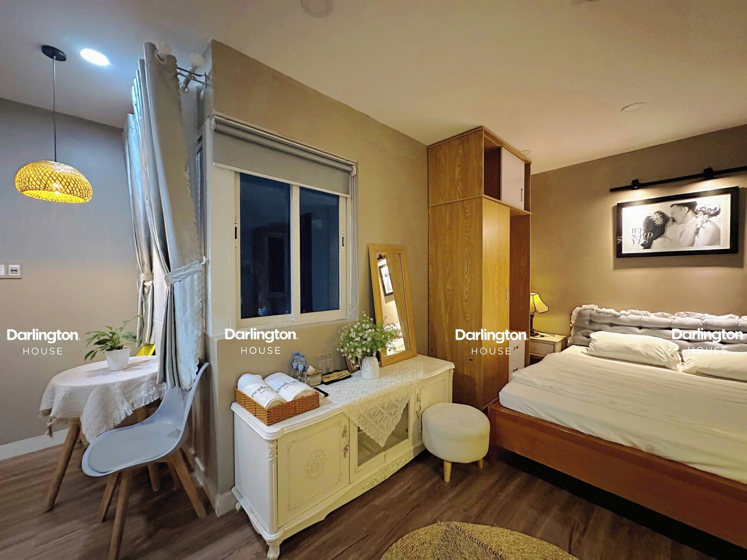 A bedroom with a bed, a vanity with a mirror, a bedside table, a window with curtains, and a hanging light fixture.