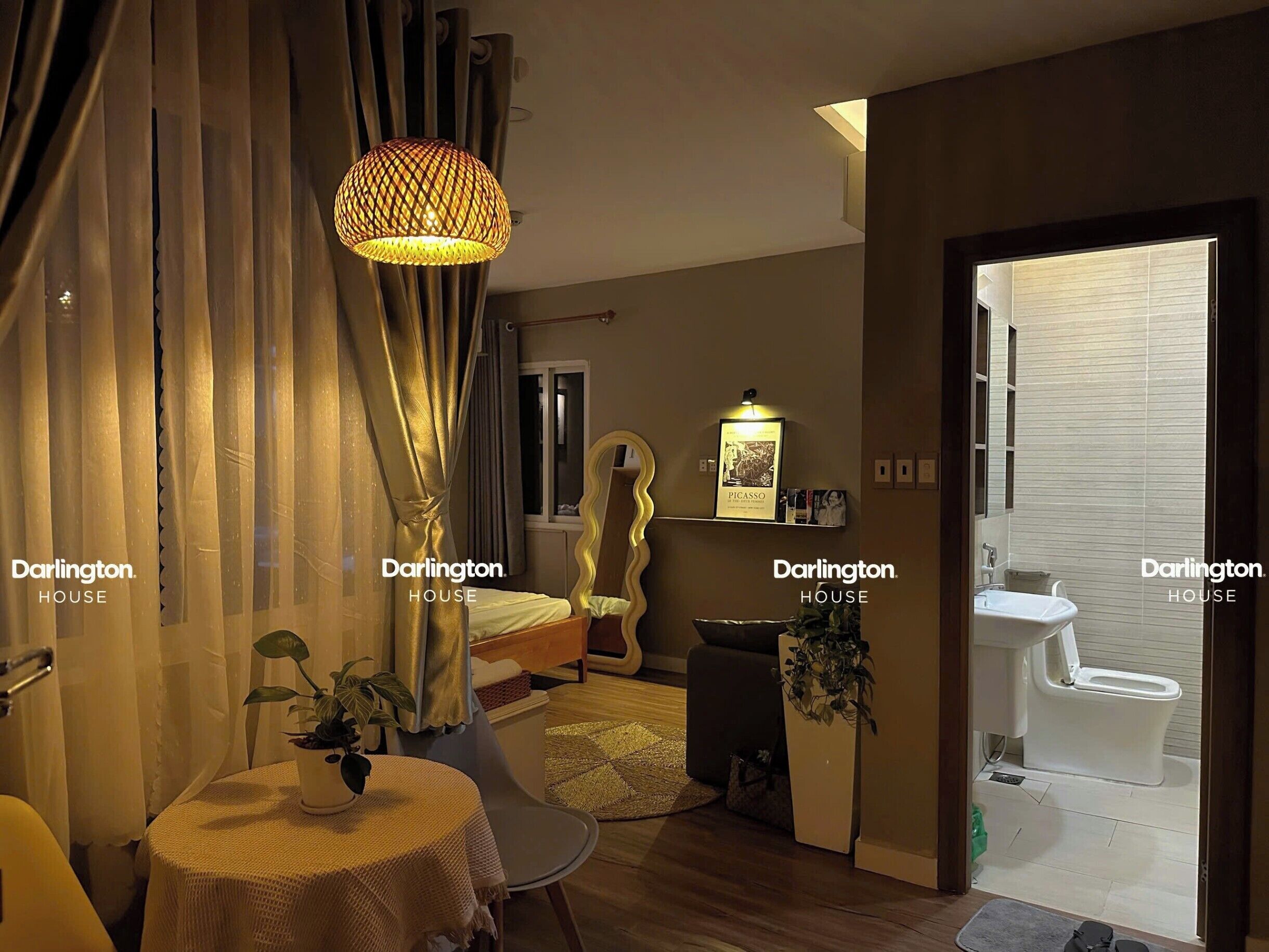 A hotel room with a bed, a bedside table with a plant, a lamp, a mirror, a bathroom with a toilet and sink, and a door leading to another room.