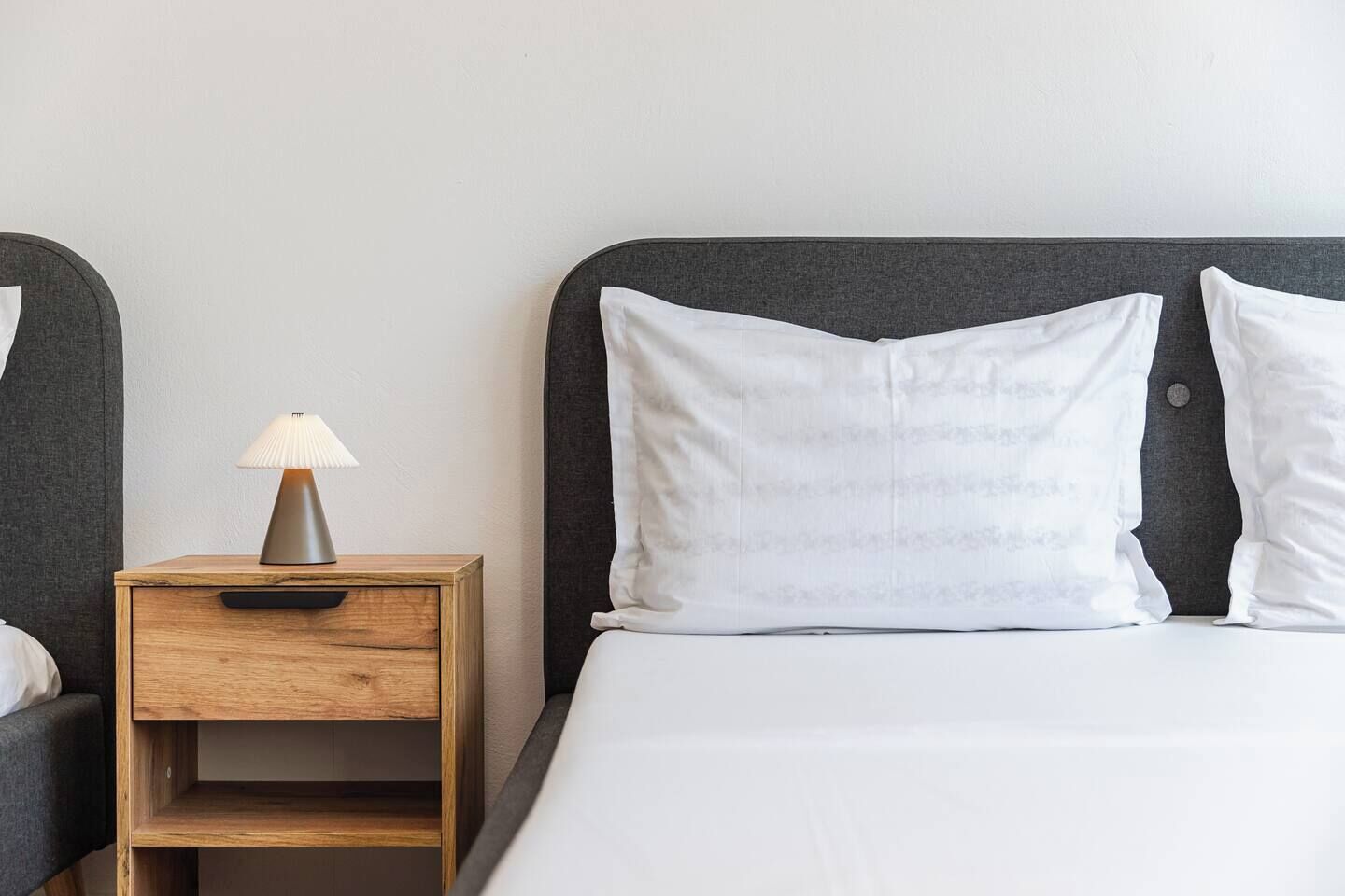 A neatly made bed with white bedding and two pillows, a bedside table with a lamp, and a wooden nightstand.