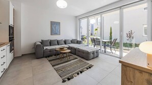 Un salon moderne comprenant un canapé d’angle, une table à manger avec des chaises, un coin cuisine avec des placards et une grande porte-fenêtre coulissante donnant sur une terrasse extérieure.