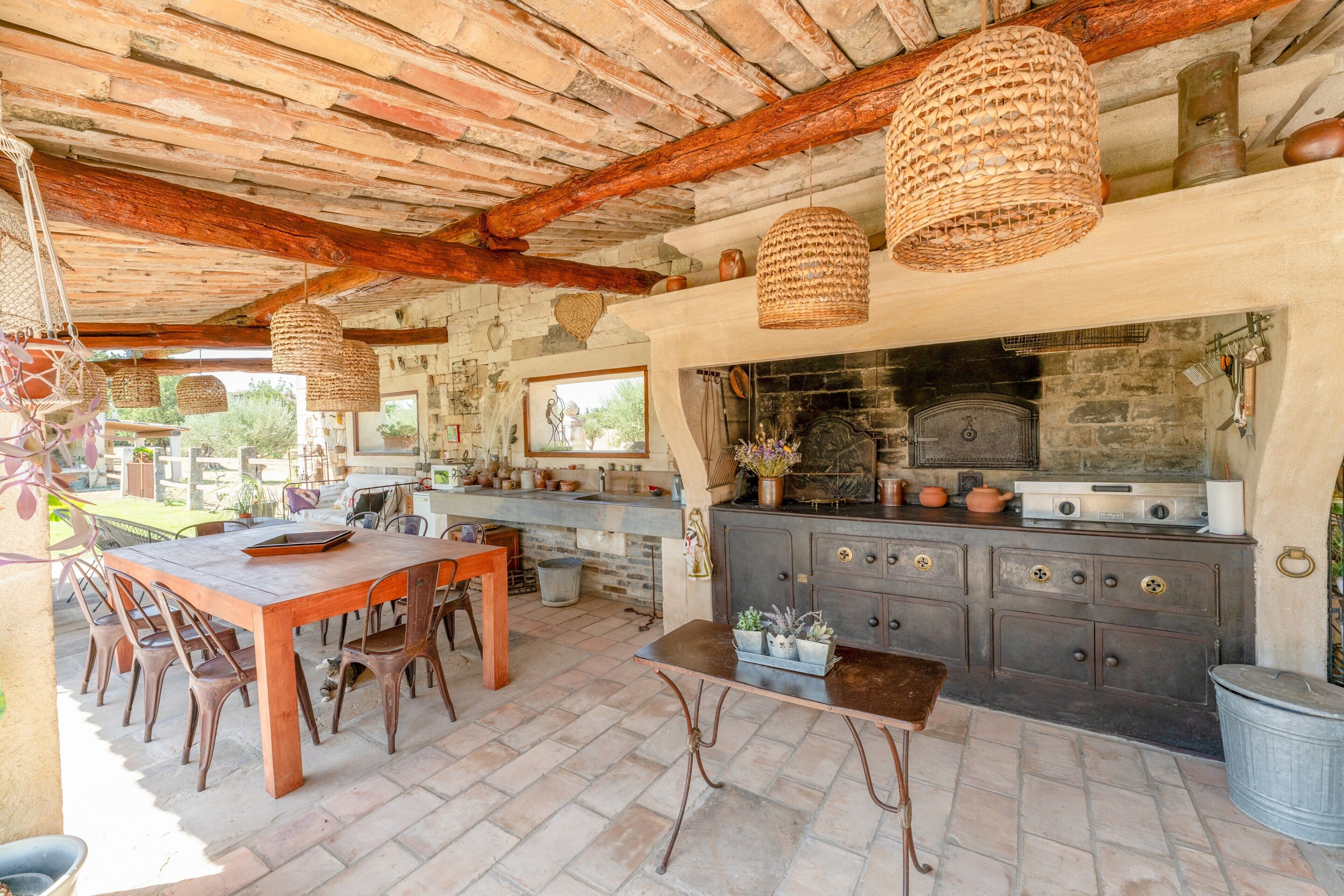 Une cuisine extérieure dotée d’une cheminée en pierre, d’une table et de chaises en bois, de luminaires suspendus en osier et d’un mur en pierre rustique.