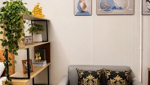 A modern living room with a grey sofa, two decorative pillows, a wooden side table, a bookshelf with plants, and wall art. - Near BKK Homestay Airport (Bang Phli)