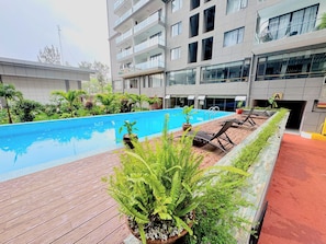 Una piscina con sillones de descanso y plantas en macetas, rodeada por un edificio de varios pisos con balcones.