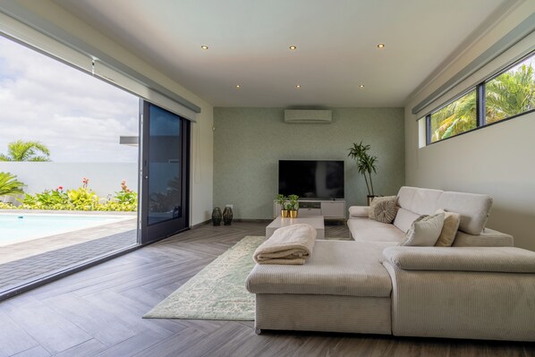Ein modernes Wohnzimmer mit einem großen Flachbildfernseher, einem beige gefärbten Sofa und Blick auf einen Swimmingpool.