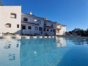 Un grand bâtiment avec une piscine devant, doté de nombreux balcons et surplombant un ciel d’un bleu éclatant.