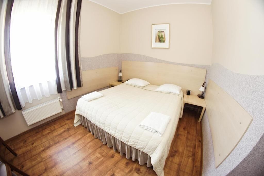 Ein Schlafzimmer mit einem großen Bett, zwei Nachttischen, einem Beistelltisch, einer Heizung und einem Fenster mit Vorhängen.