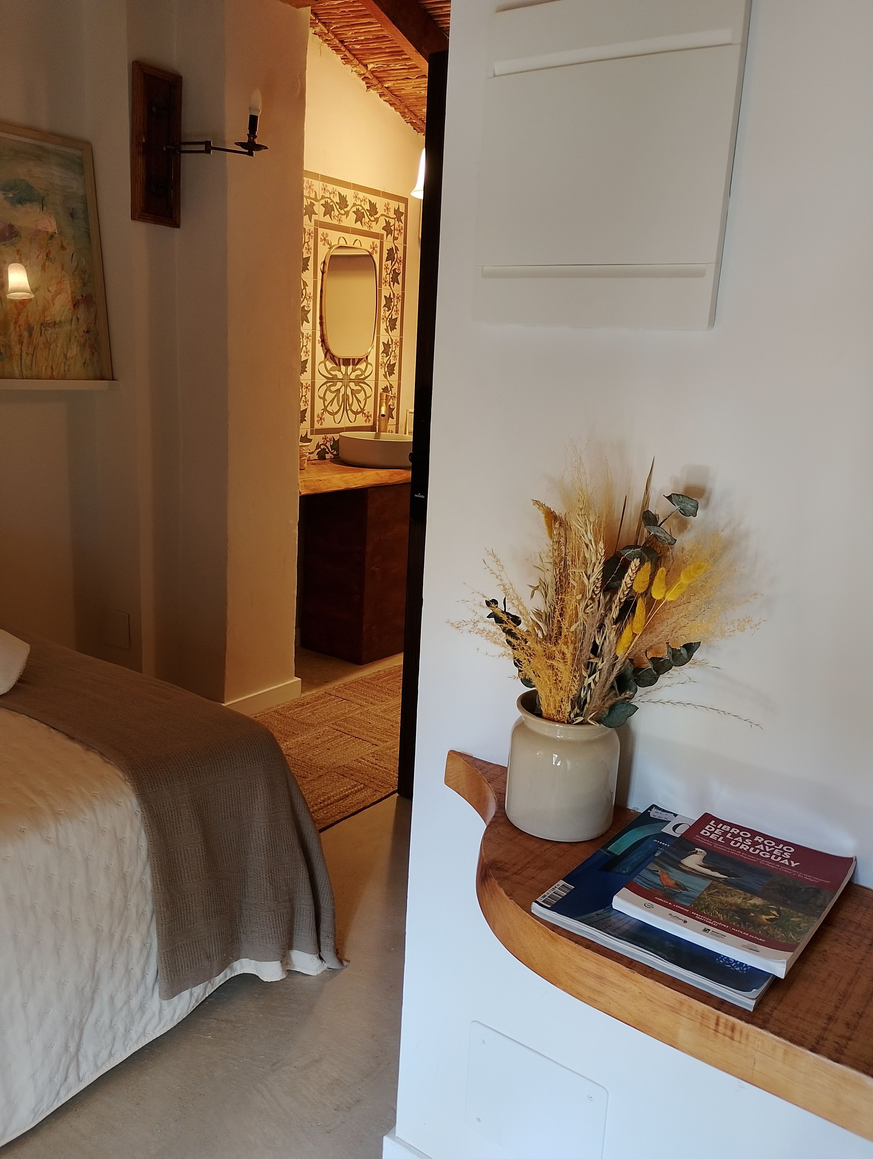 Un dormitorio con cama, una mesita de noche con un libro y un jarrón de plantas secas, y un baño con espejo y paredes con azulejos.