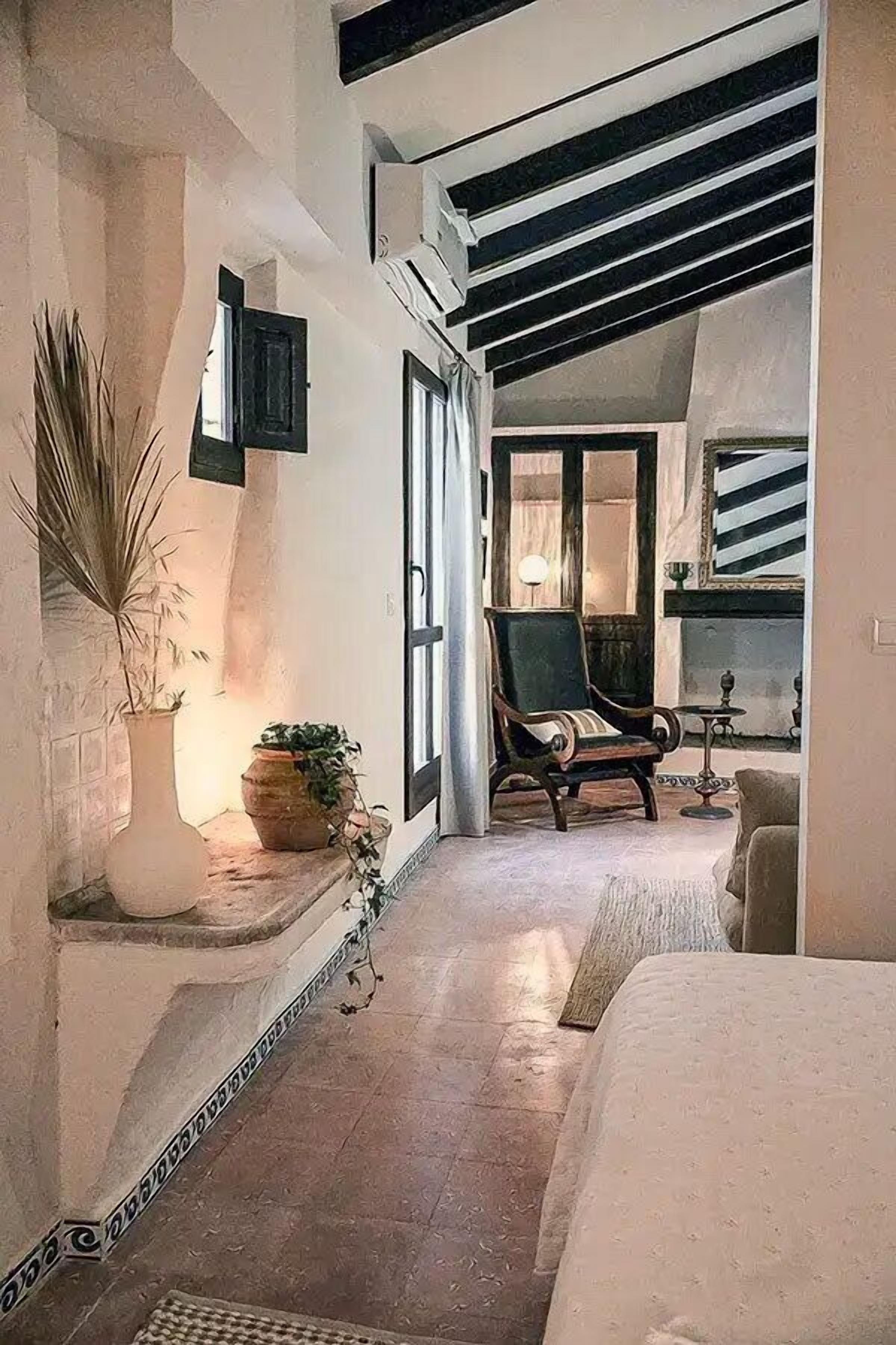 Un interior acogedor con una cama, una silla y una pequeña mesa. Hay una planta en maceta y un jarrón decorativo en un estante.