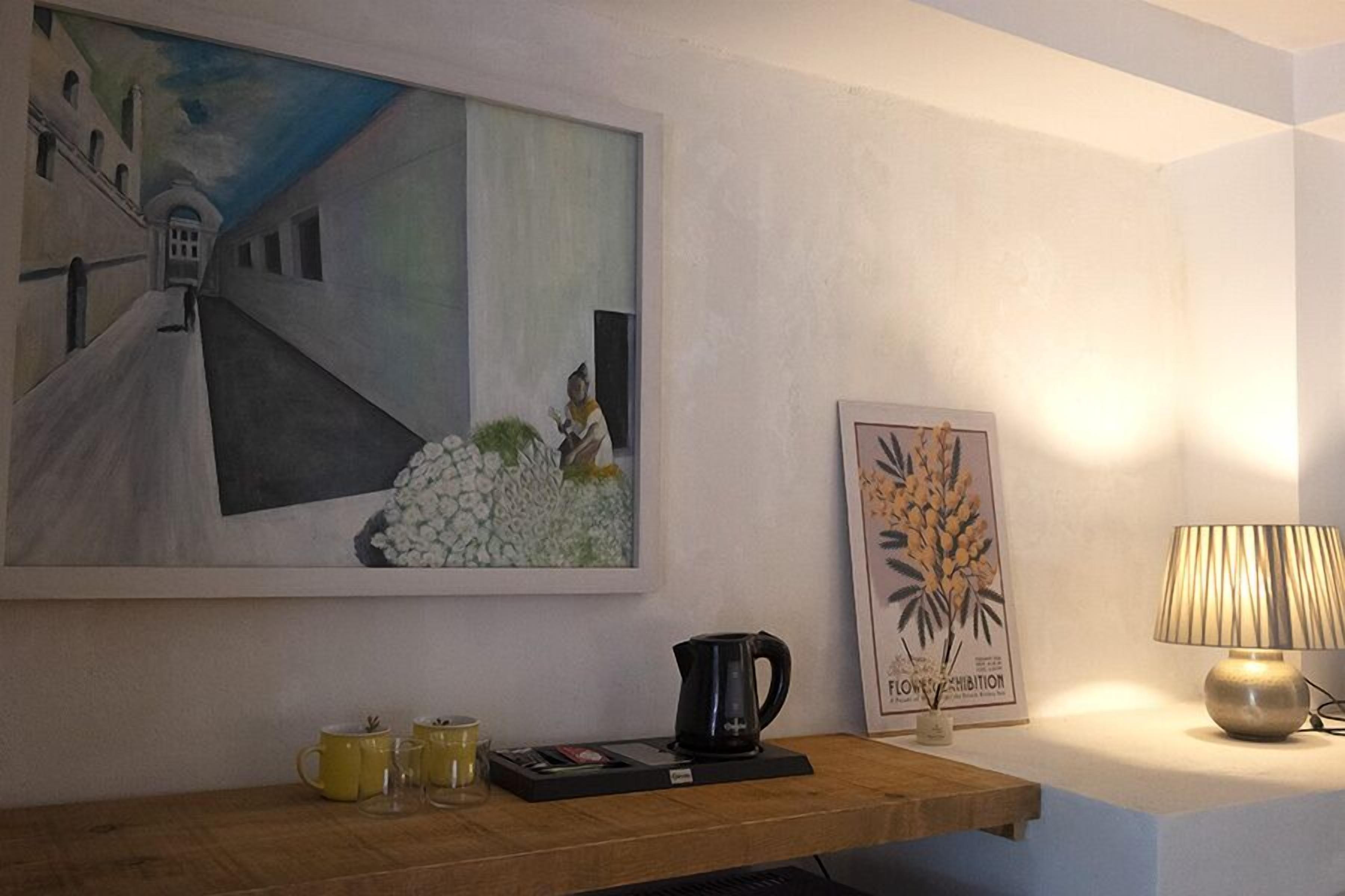 Une chambre avec un tableau, une estampe botanique encadrée, une machine à café et une lampe sur une table de chevet.