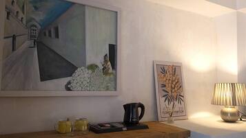 Une chambre avec un tableau, une estampe botanique encadrée, une machine à café et une lampe sur une table de chevet.