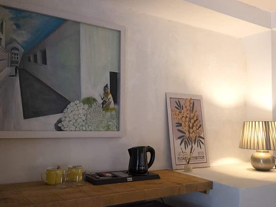 A room with a painting, a framed botanical print, a coffee maker, and a lamp on a bedside table.