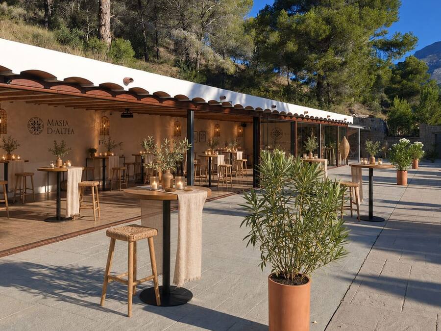 Un espacio de comedor al aire libre con mesas y sillas, rodeado de macetas con plantas y un paisaje montañoso al fondo.