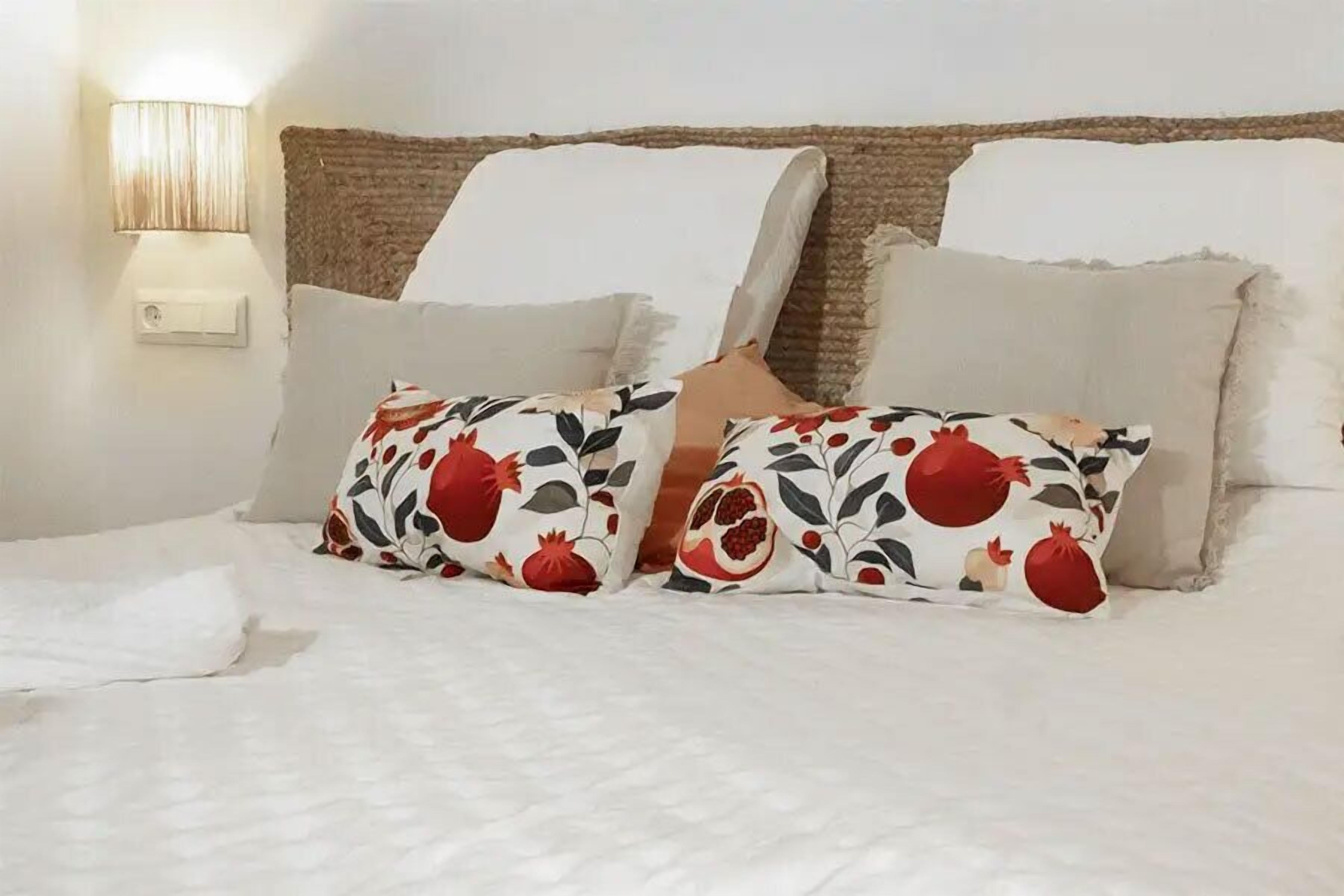 Una cama con ropa de cama blanca, dos almohadas decorativas con estampado floral y una lámpara de pared.