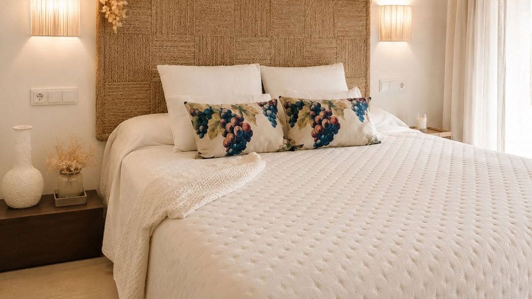 Una cama bien hecha con un edredón blanco texturizado, dos almohadas decorativas con diseño de uvas, un cabecero tejido y apliques de pared.
