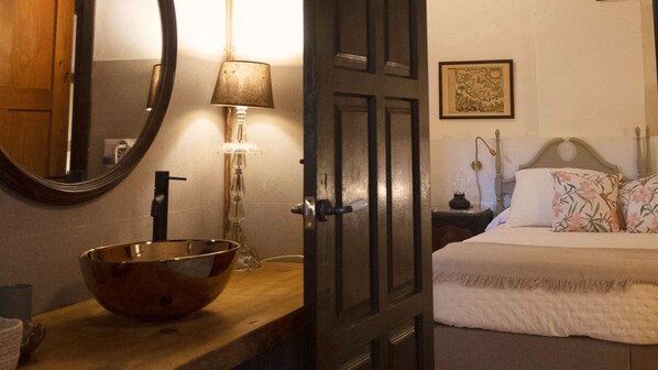 A hotel room with a wooden vanity, a bowl sink, a mirror, a bedside lamp, a bed with floral pillows, and a framed picture on the wall.