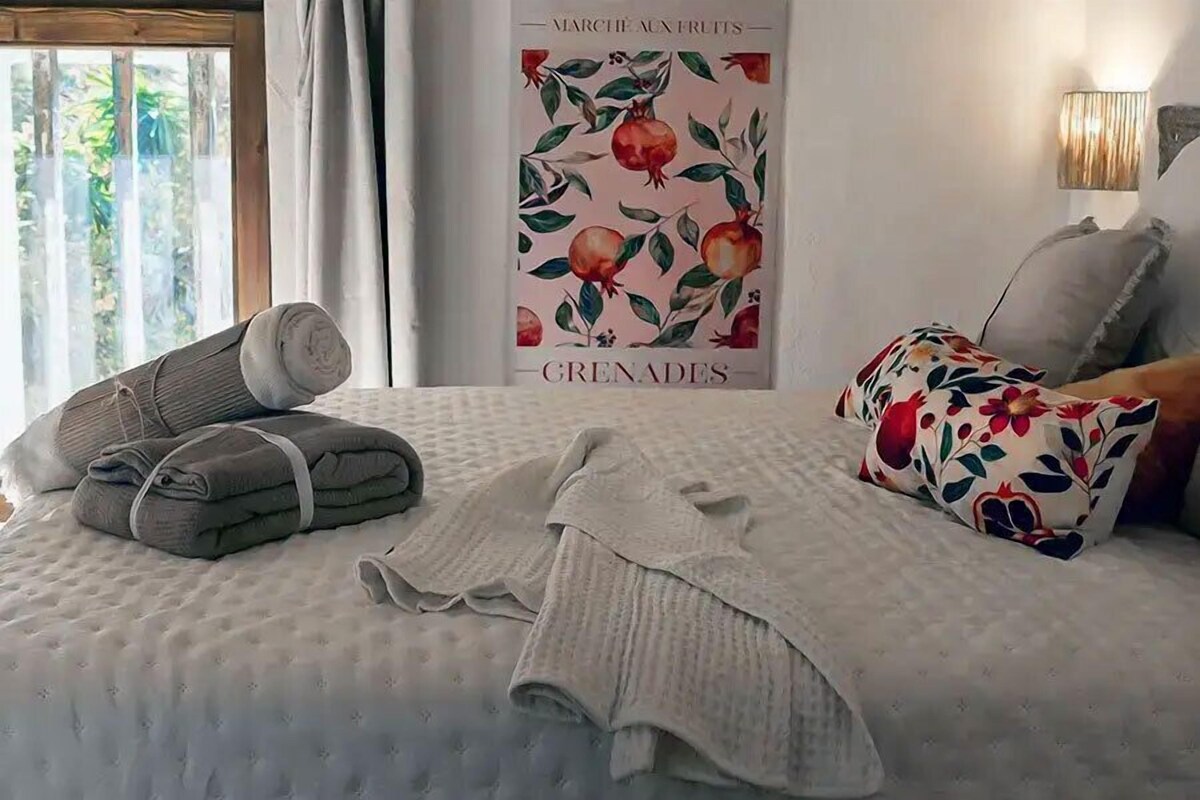 Una cama con cubre cama blanco, un cartel de granadas y una ventana con cortinas.