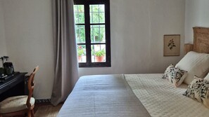 A bedroom with a bed, a desk, a chair, a window with a view of greenery, and a framed picture on the wall.