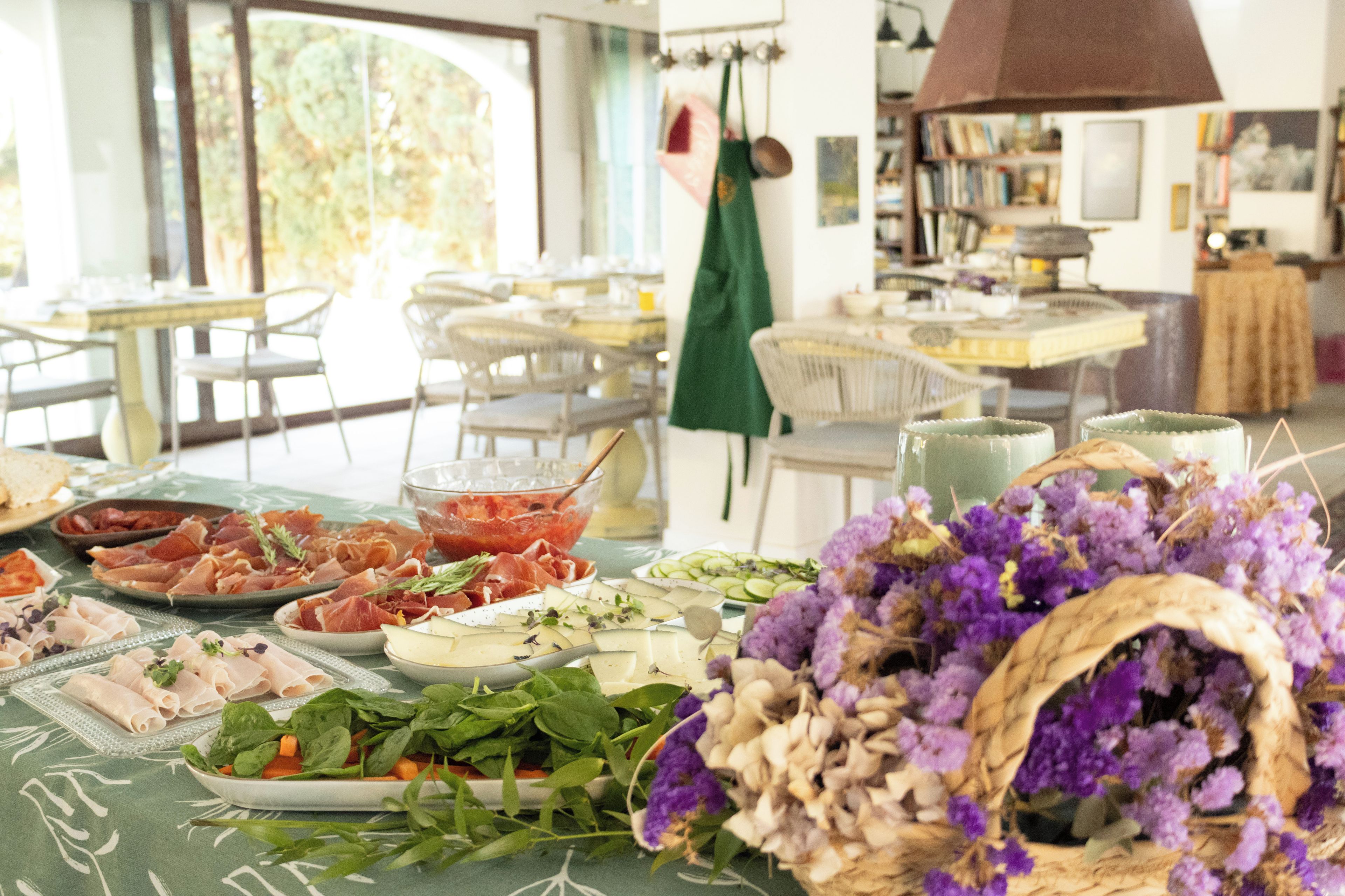 A dining area with a table set for a meal, featuring various food items and a basket of purple flowers.