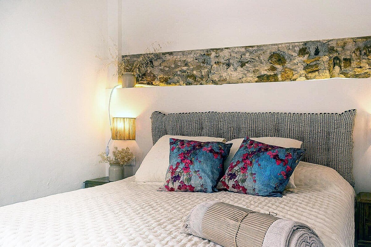 Una cama con edredón blanco texturizado, dos almohadas decorativas con motivos florales, un cabecero con diseño texturizado y una lámpara de pared sobre una mesita de noche.